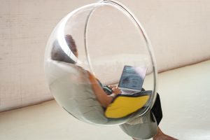 A person sitting inside a clear, bubble-like chair, using a laptop with a yellow laptop sleeve next to them.