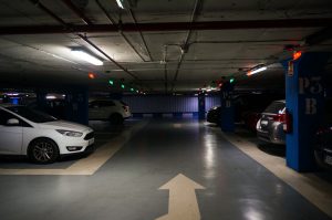 A dimly lit underground parking garage with several cars parked along the sides. Blue columns separate the parking spaces. The ceiling has colored lights above parking spots indicate availability. An arrow is painted on the floor pointing forward.