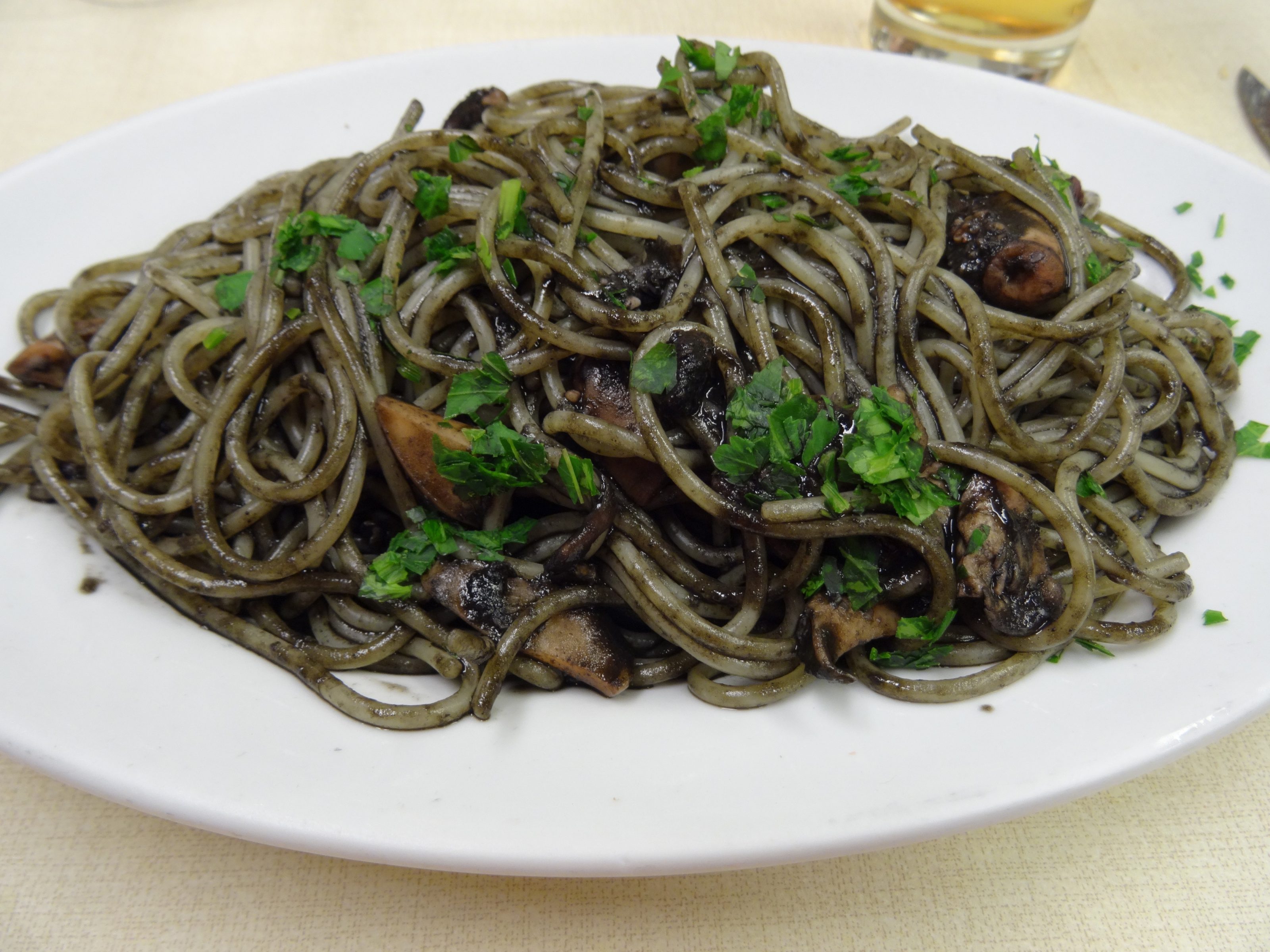 A plate of spaghetti al nero di seppia coated in black squid ink sauce, garnished with chopped parsley and mixed with pieces of seafood, served on a white plate.