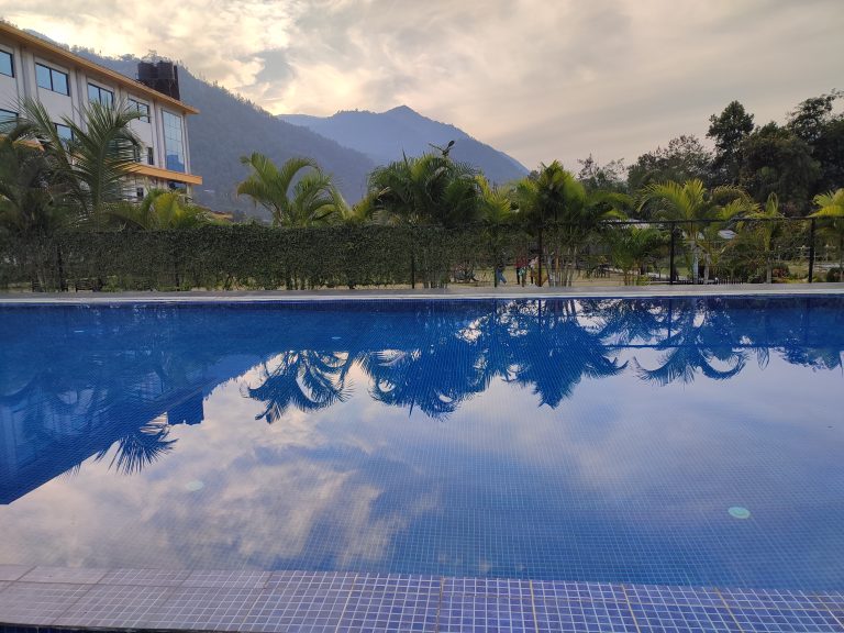 A serene outdoor swimming pool reflects the cloudy sky and palm trees. In the background, there is a landscape of lush green trees and hills, with part of a building visible on the left side.