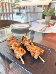 Grilled chicken skewers on a black rectangular plate, placed on a glass table with a blurred background of an outdoor dining area.