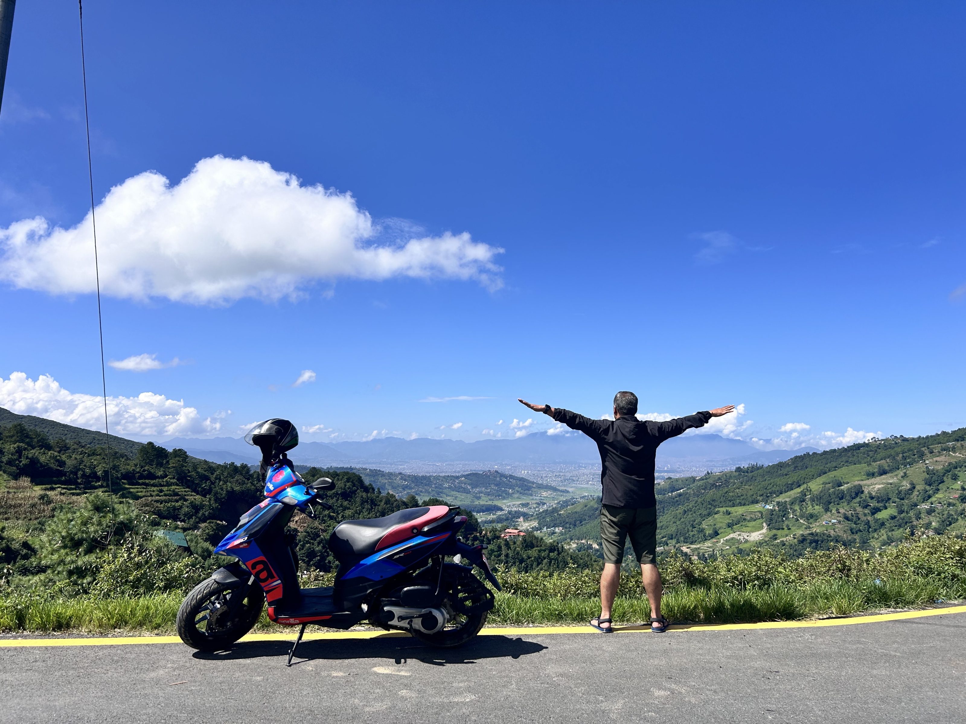 A person with outstretched arms stands besinde a motorcycle on a roadside overlooking a scenic valley, with green hills and a city in the distance under a clear blue sky.