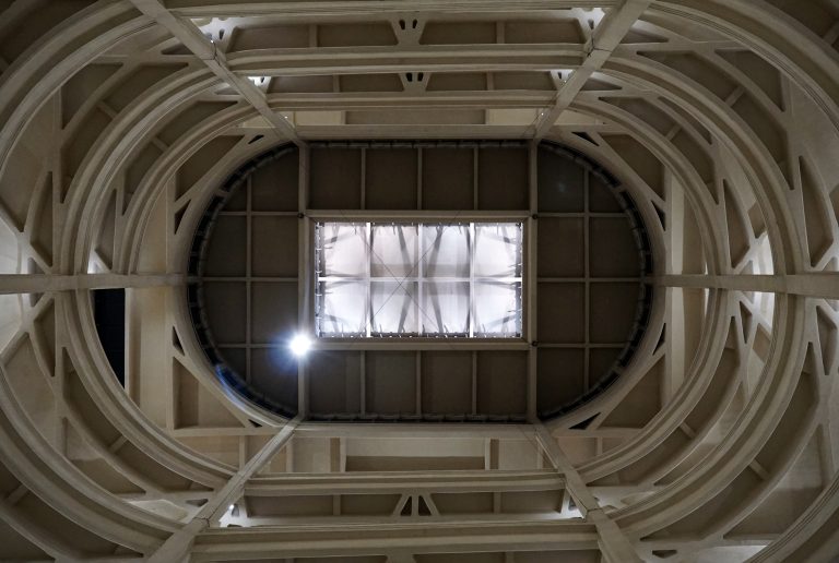 Looking up at a geometric ceiling with concentric circular patterns and a rectangular skylight at the center inside the Lingotto building. The structure features curved arches and parallel beams, creating a modern architectural design with a central light source illuminating the scene.