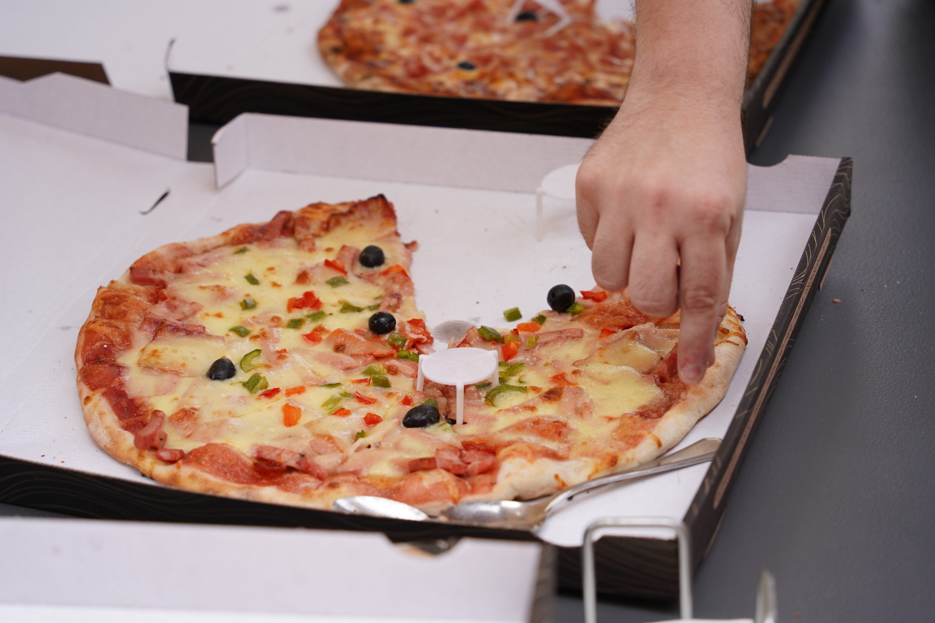 A person reaching for a slice of pizza in a cardboard box. The pizza is topped with cheese, black olives, red and green bell peppers, and pieces of bacon. Another pizza is partially visible in the background