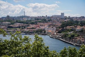 A scenic view of a Porto, Portugal with Douro river in the foreground, lined with buildings featuring red-tiled roofs. Modern and historical buildings are visible in the background, along with a television tower on a hill.