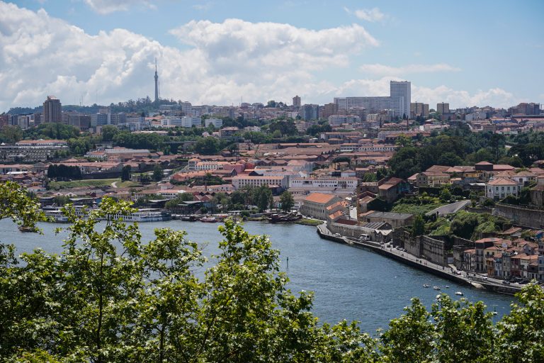 A scenic view of a Porto, Portugal with Douro river in the foreground, lined with buildings featuring red-tiled roofs. Modern and historical buildings are visible in the background, along with a television tower on a hill.