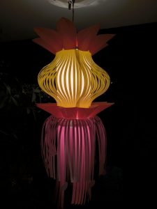 A brightly lit, multi-colored paper lantern hanging against a dark background. The lantern features a yellow bulbous central section with red petal-like extensions at the top and pink fringe details hanging below.
