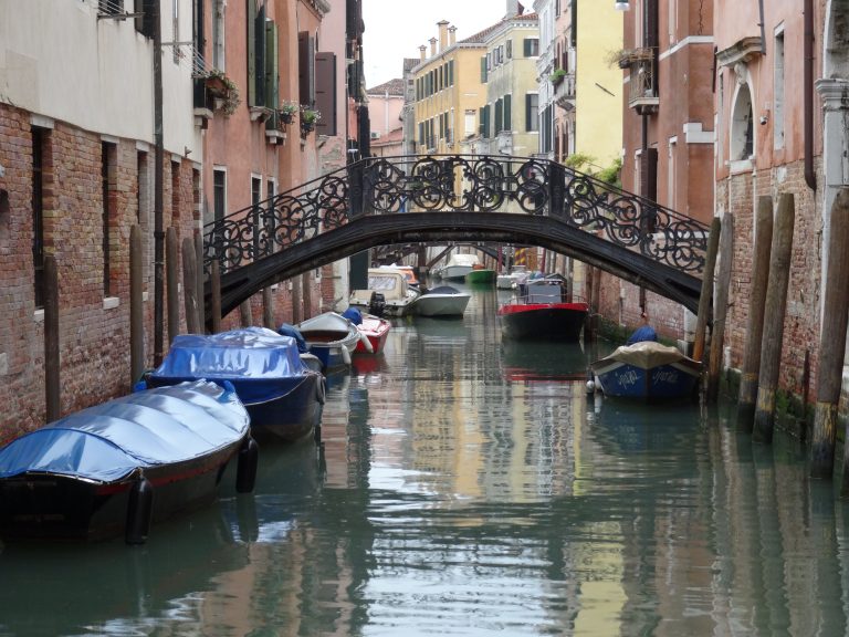 A picturesque canal in Venice with several boats moored along the sides. A decorative iron bridge arches over the canal, surrounded by traditional buildings with colorful facades and window boxes with flowers.