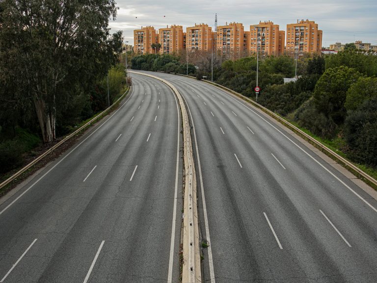 A wide, empty highway with three lanes in each direction, separated by a concrete median. The road is surrounded by trees and greenery on both sides, and in the distance, there are several orange-colored apartment buildings under an overcast sky.