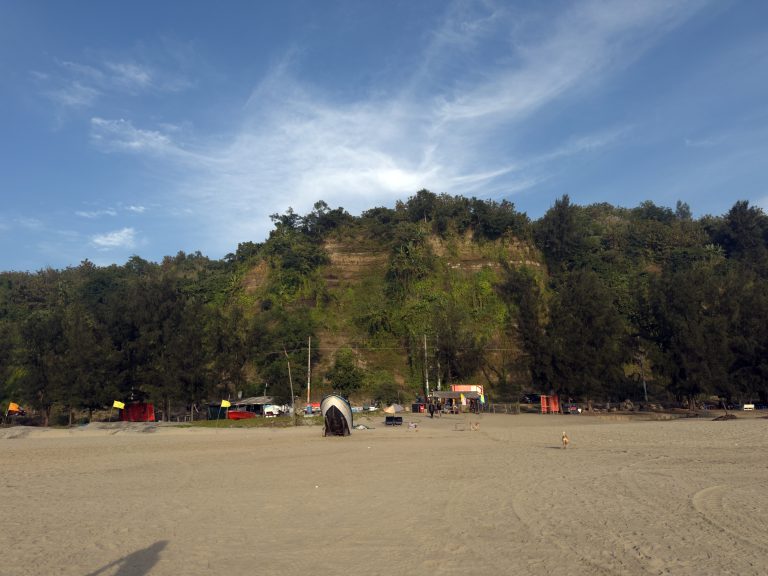 A beach with a wide expanse of sand in the foreground, bordered by a dense, green hill in the background. There is a small boat on the sand, and some colorful stalls are visible near the base of the hill.