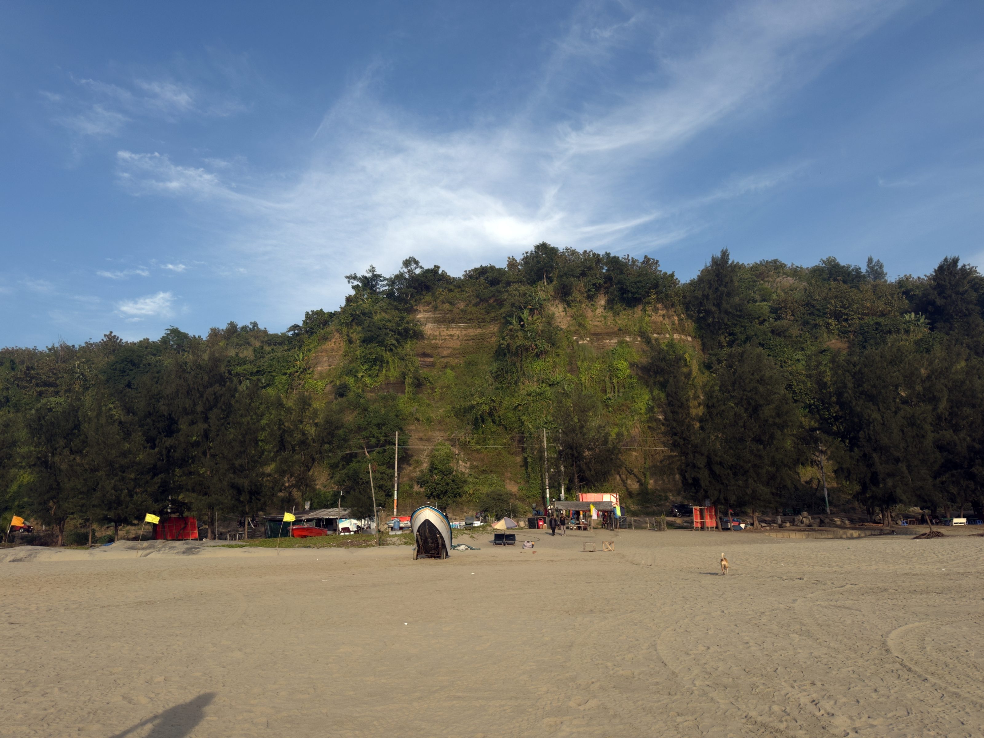 A beach with a wide expanse of sand in the foreground, bordered by a dense, green hill in the background. There is a small boat on the sand, and some colorful stalls are visible near the base of the hill.