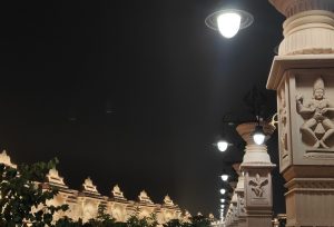 Night view of a beautifully lit architectural walkway featuring intricately carved sculptures on pillars, with glowing streetlights and greenery in the foreground.