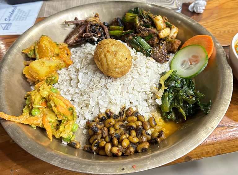 A tradition—Newari meal served on a metal plate featuring beaten rice topped with a fried ball, accompanied by various side dishes including cooked potatoes, mixed vegetables, beans, leafy greens, and garnished with cucumber and carrot slices.