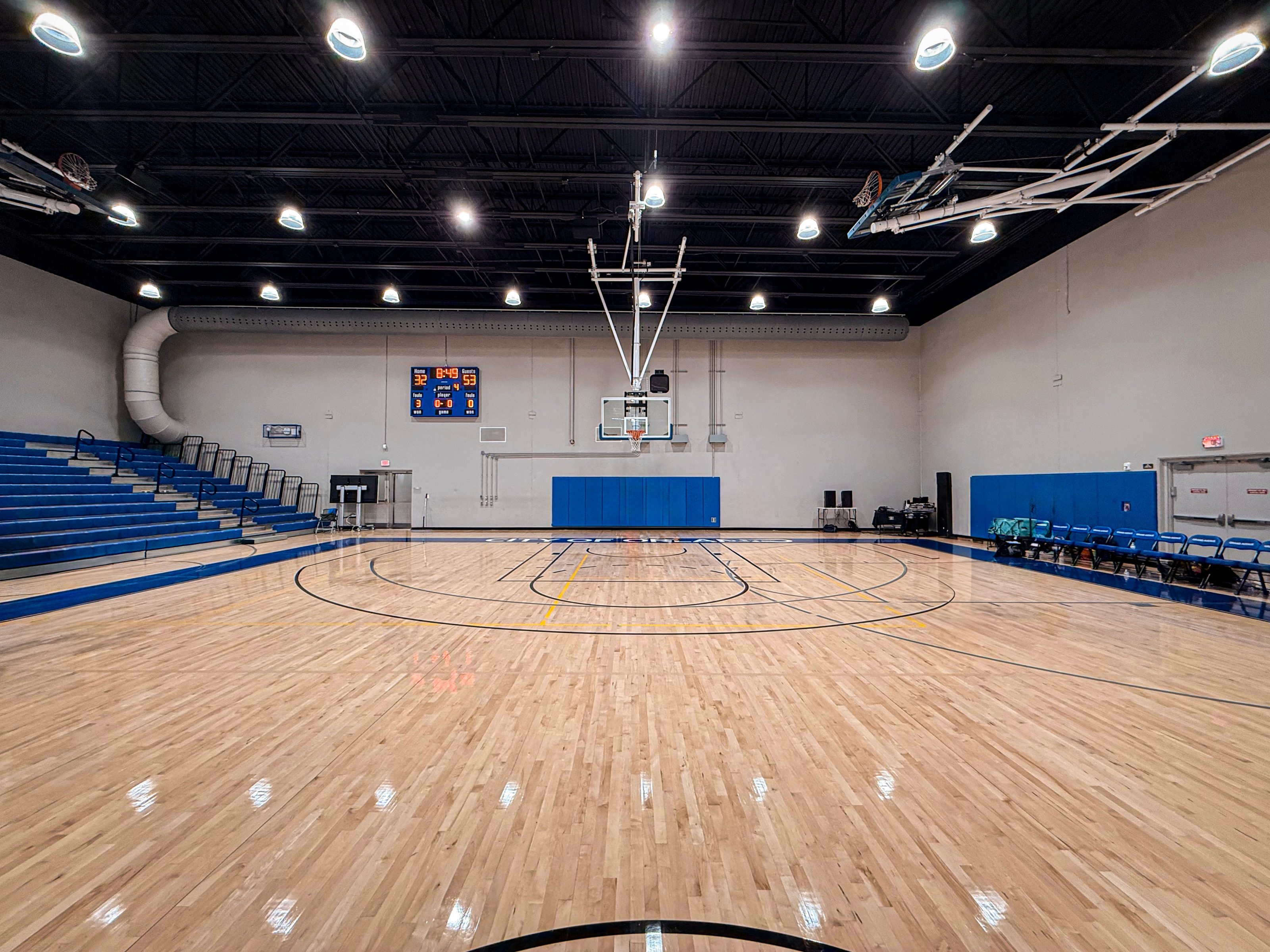 An empty indoor basketball court with polished wooden flooring, blue and white bleachers on one side, a scoreboard displaying the score, and basketball hoops on either end. The gymnasium is well-lit with overhead lighting and has blue padding on the walls.