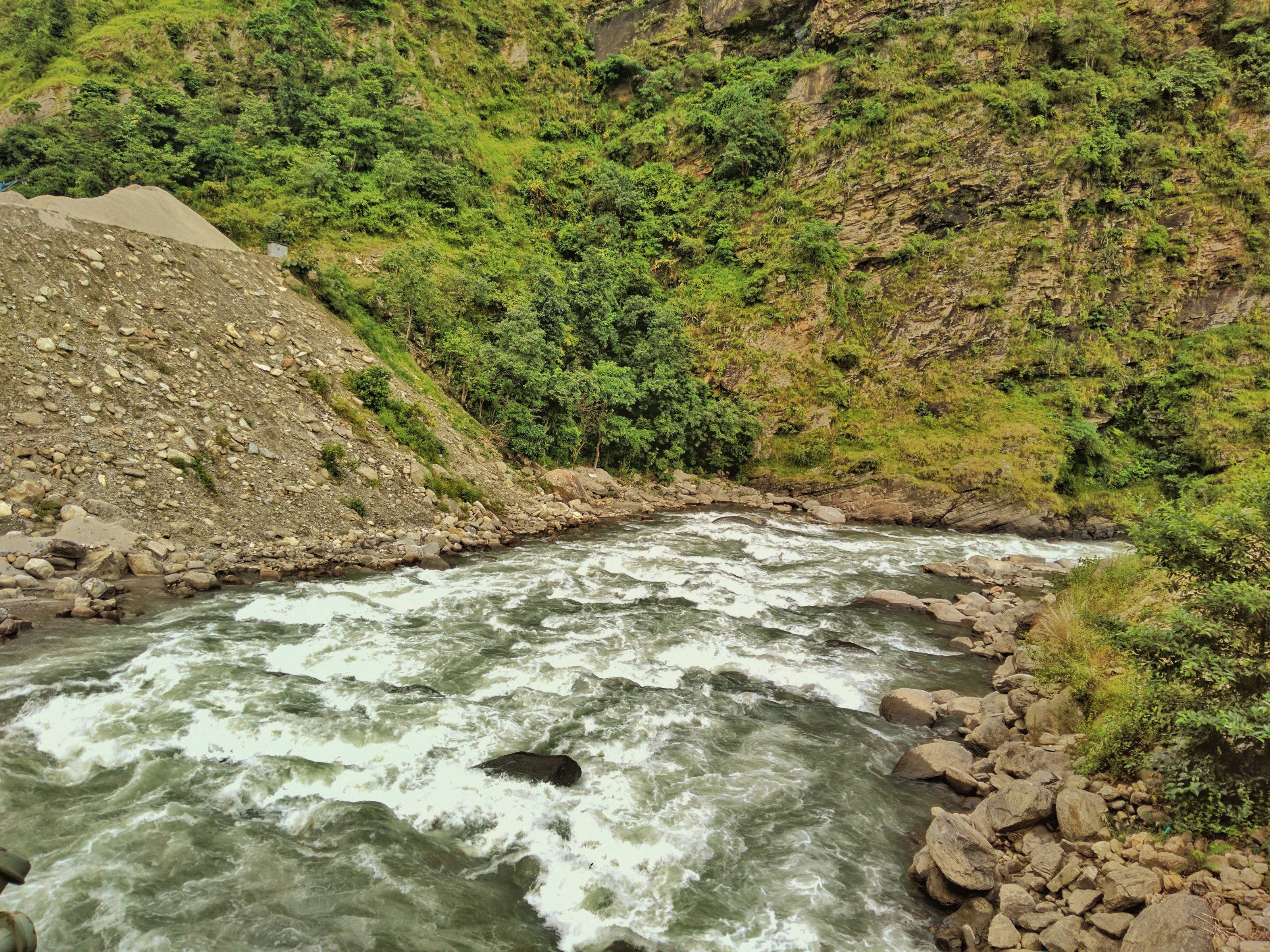 A river with white rapids flows through a rocky landscape bordered by lush green hills and vegetation. The riverbank is dotted with large rocks and a section of loose stones on the left side.