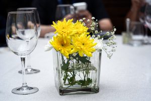 A square glass vase containing yellow and white flowers, placed on a table with empty wine glasses nearby. There are blurred people in the background.