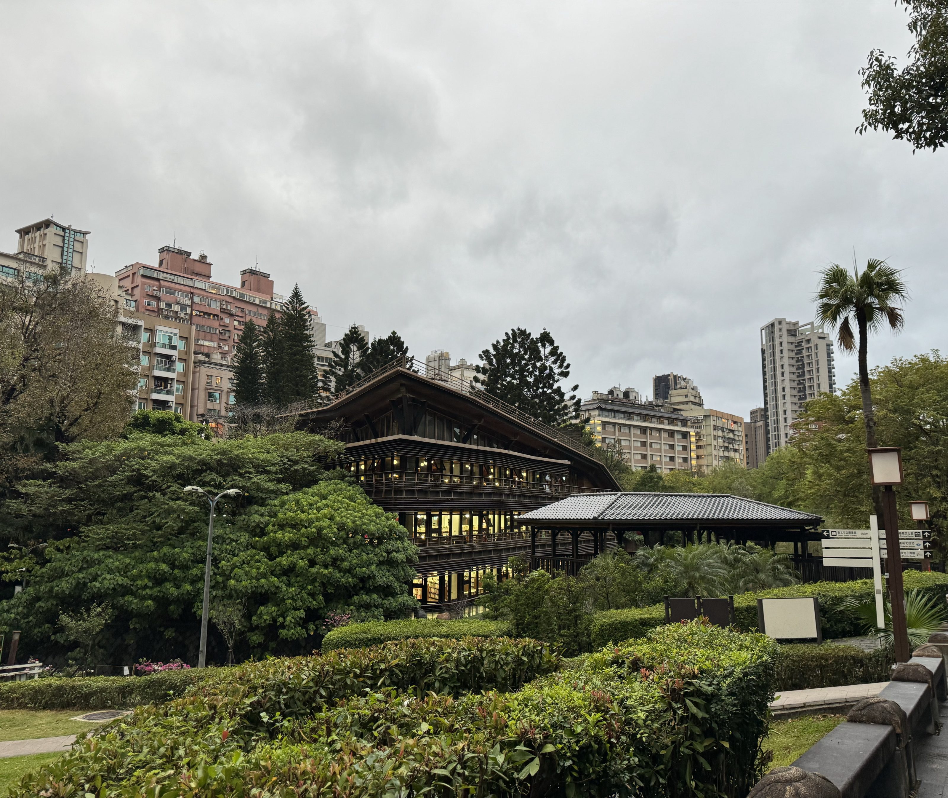 A modern wooden building surrounded by lush greenery and trees, set against a backdrop of urban apartment buildings and a cloudy sky. A well-maintained garden with trimmed hedges and a palm tree is visible in the foreground.