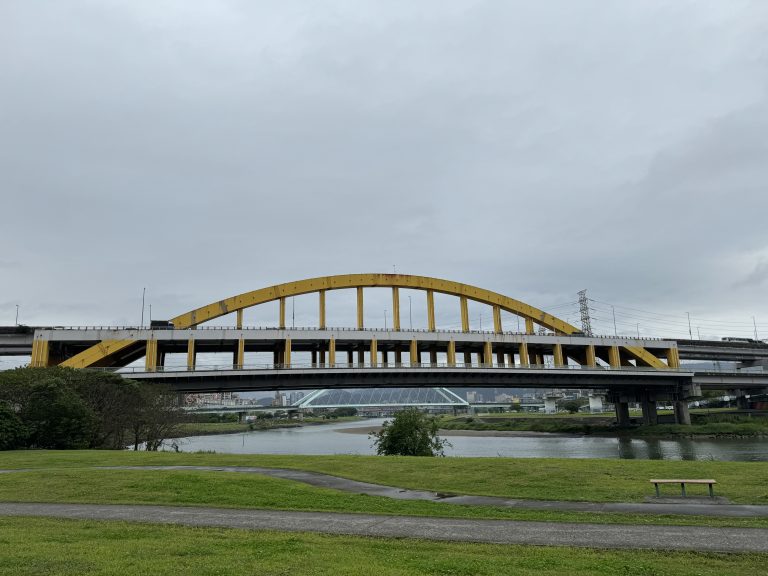 A large yellow arch bridge spans over a river on a cloudy day. The foreground features a grassy area with a paved path and a wooden bench. Buildings and another bridge can be seen in the distance.