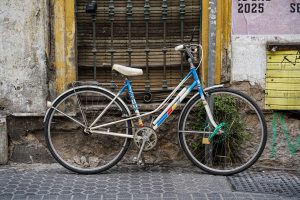 A vintage-style bicycle with a white and blue frame is parked against a worn, old building with peeling paint and a shuttered window. The bike has a small padlock securing it to a nearby pole, and there are faded posters and graffiti on the wall. 