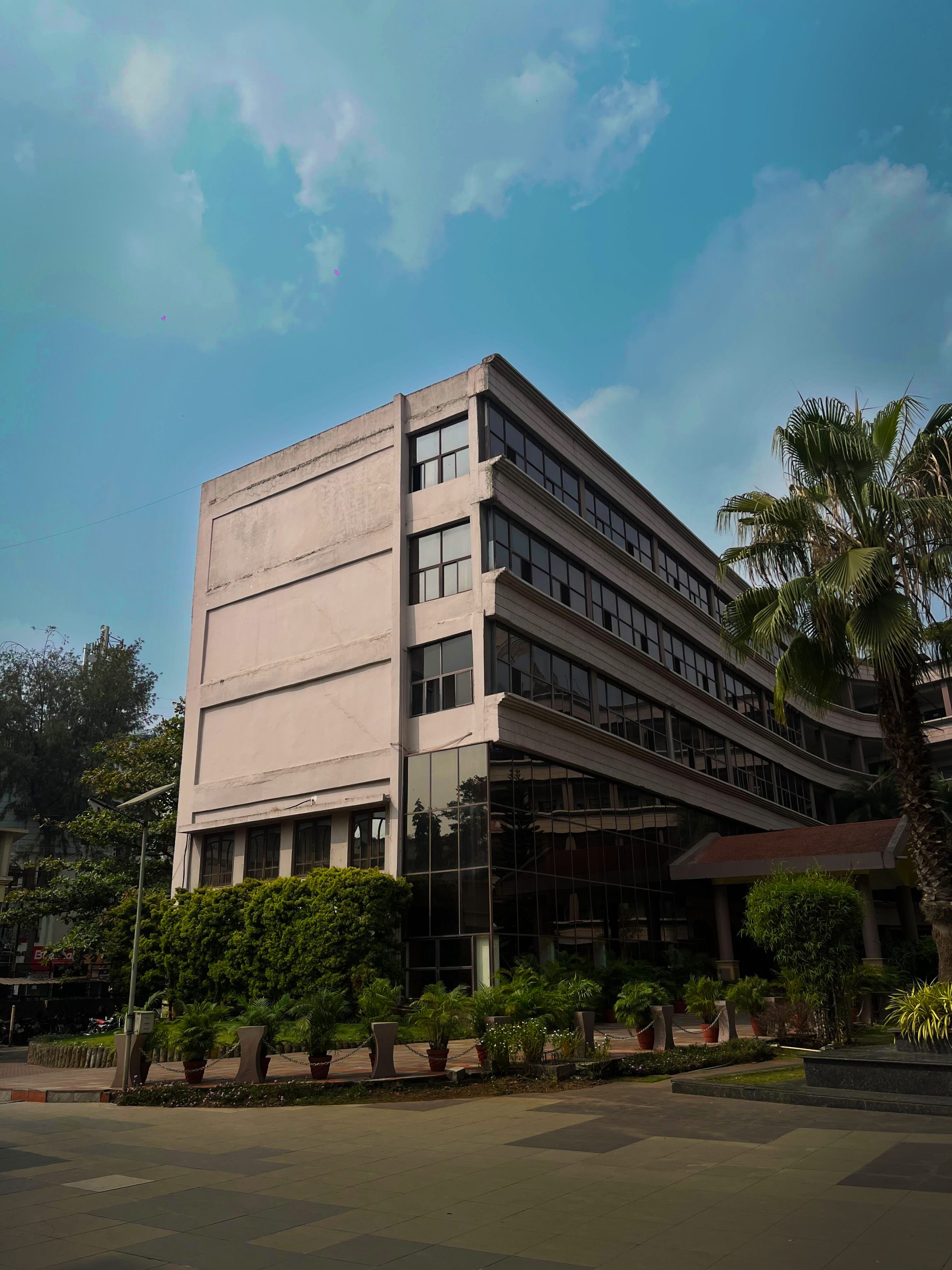 A modern multi-story building with large glass windows, clean lines, and lush greenery, including potted shrubs and a tall palm tree, set against a bright, cloudy sky.