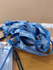 A close-up of a pile of blue lanyards on a wooden surface, featuring the WordPress logo and text. A pen and a clear plastic badge holder are also visible.