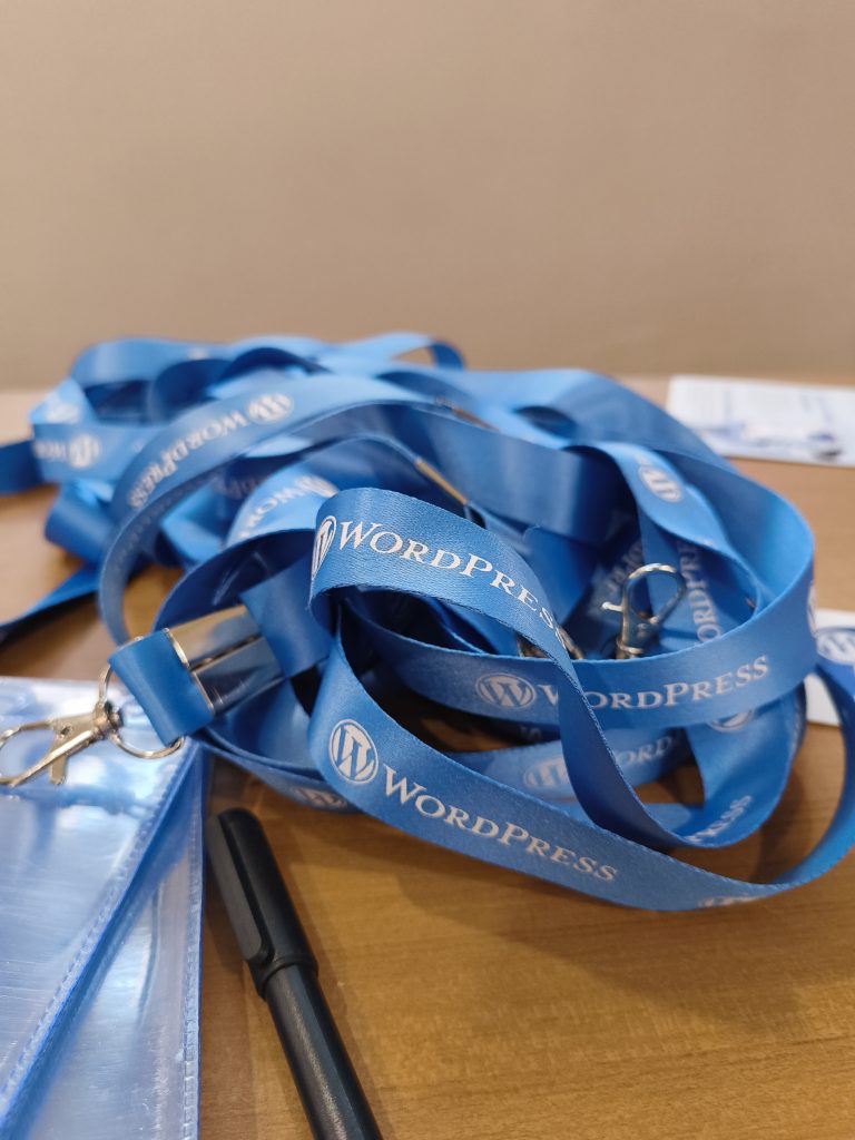 A close-up of a pile of blue lanyards on a wooden surface, featuring the WordPress logo and text. A pen and a clear plastic badge holder are also visible.