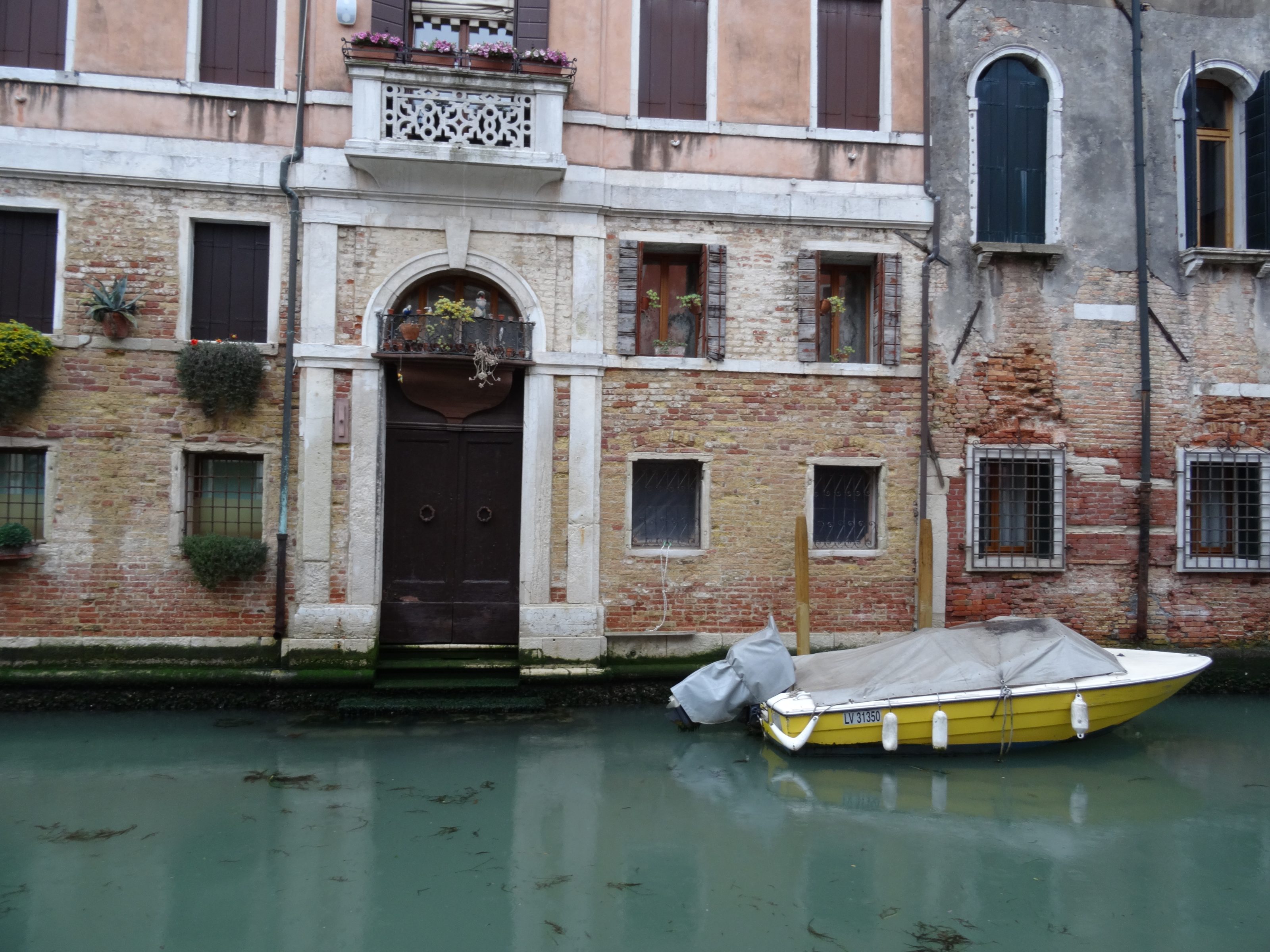 A picturesque canal scene featuring a weathered brick building with arched windows and a decorative balcony. In front of the building, a yellow boat with a gray cover is moored in the greenish water. There are small plants and greenery decorating the facade.