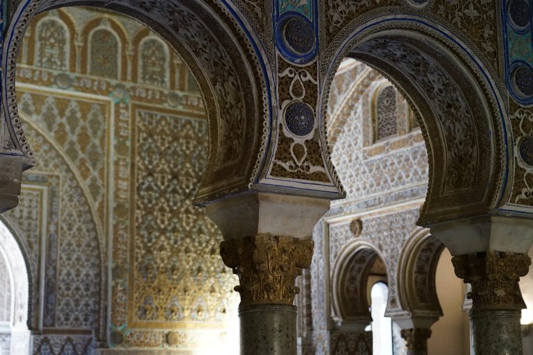 Ornate interior of a historic building featuring intricate Islamic architectural designs. The image shows detailed arches with elaborate carvings and patterns in shades of blue, gold, and brown, supported by decorated columns.