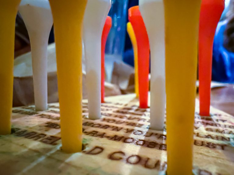 Close-up view of colorful golf tees inserted into holes on a wooden board with text partially visible on the surface.