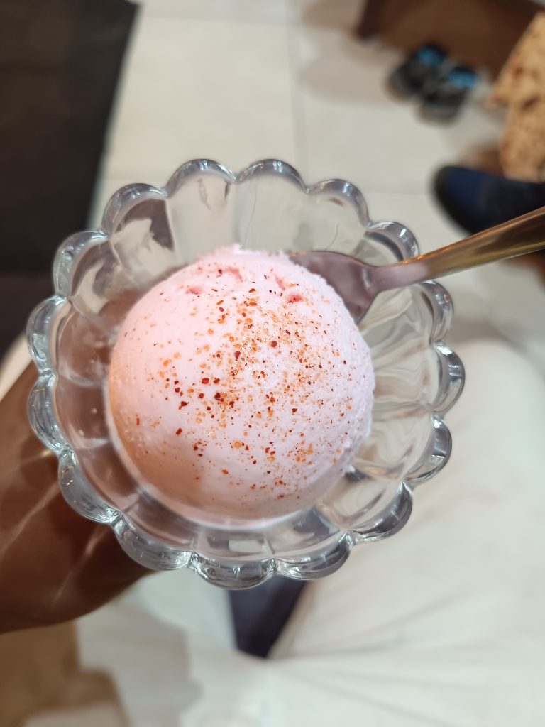 A close-up of a scoop of pink ice cream in a glass bowl, sprinkled with red chili powder. A spoon is placed inside the bowl. The background shows a tiled floor and indistinct objects.