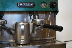 Close-up of a professional espresso machine, showing the portafilter, steam wand, and a stainless steel milk frothing pitcher. The machine's control panel and temperature gauge are visible on the top.