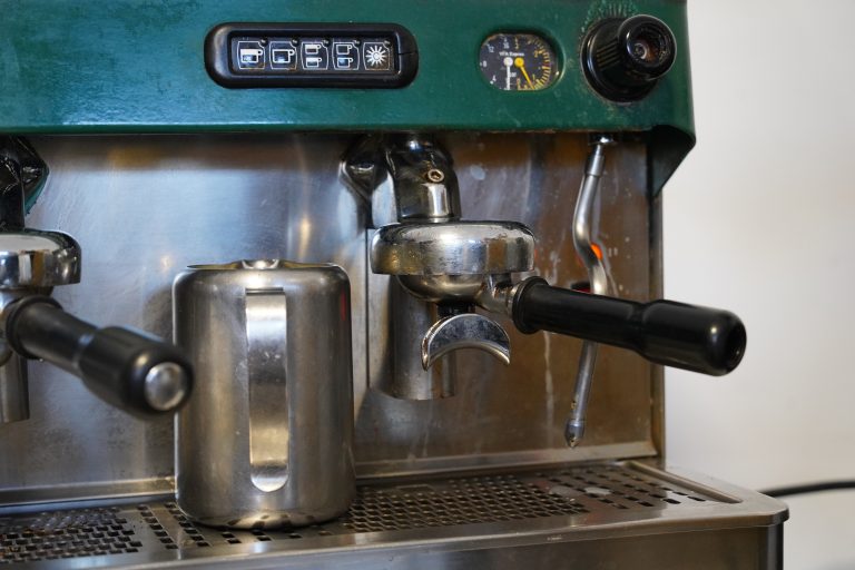 Close-up of a professional espresso machine, showing the portafilter, steam wand, and a stainless steel milk frothing pitcher. The machine’s control panel and temperature gauge are visible on the top.