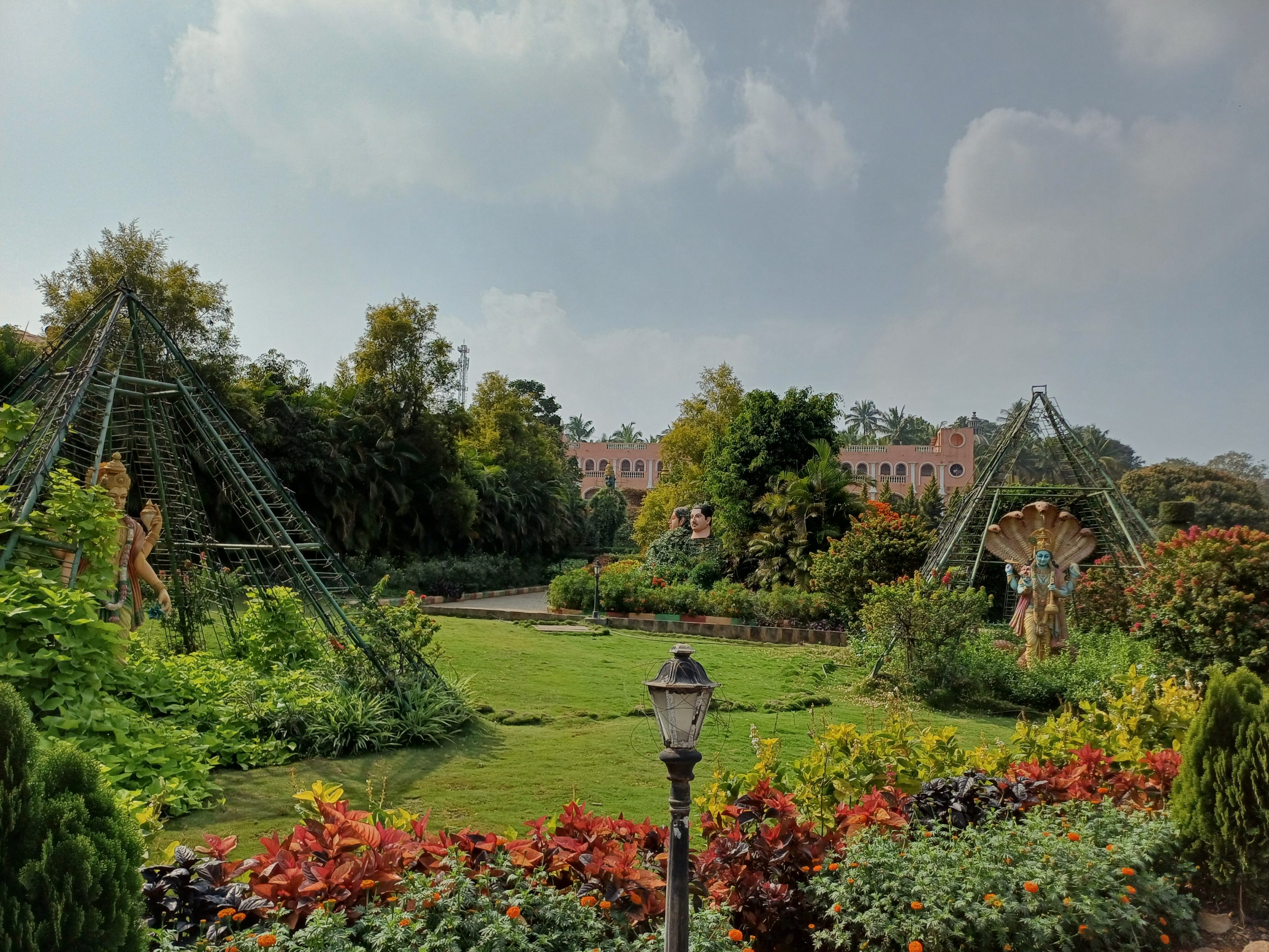 A scenic garden with vibrant flowers and greenery, featuring a historic building with arches in the background. A decorative lamppost and stone border are in the foreground, with a statue and a structure resembling a pyramid visible in the distance under a partly cloudy sky