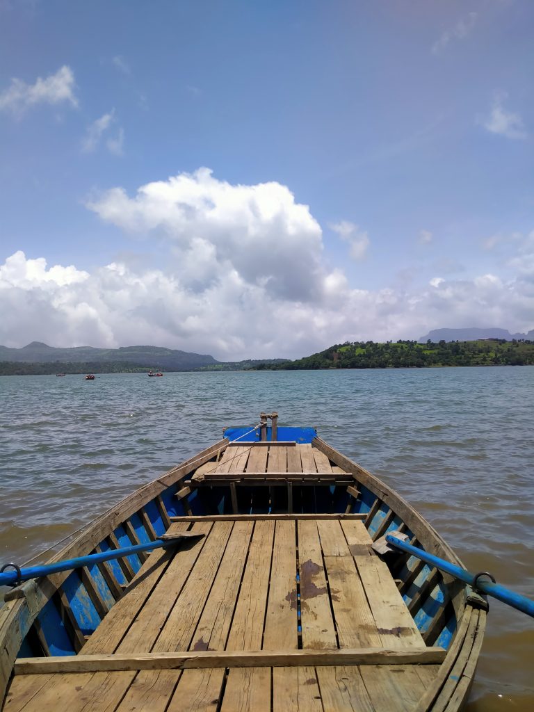 A wooden boat with blue details is floating on a calm lake. The sky is clear with a few fluffy clouds, and distant hills can be seen on the horizon.