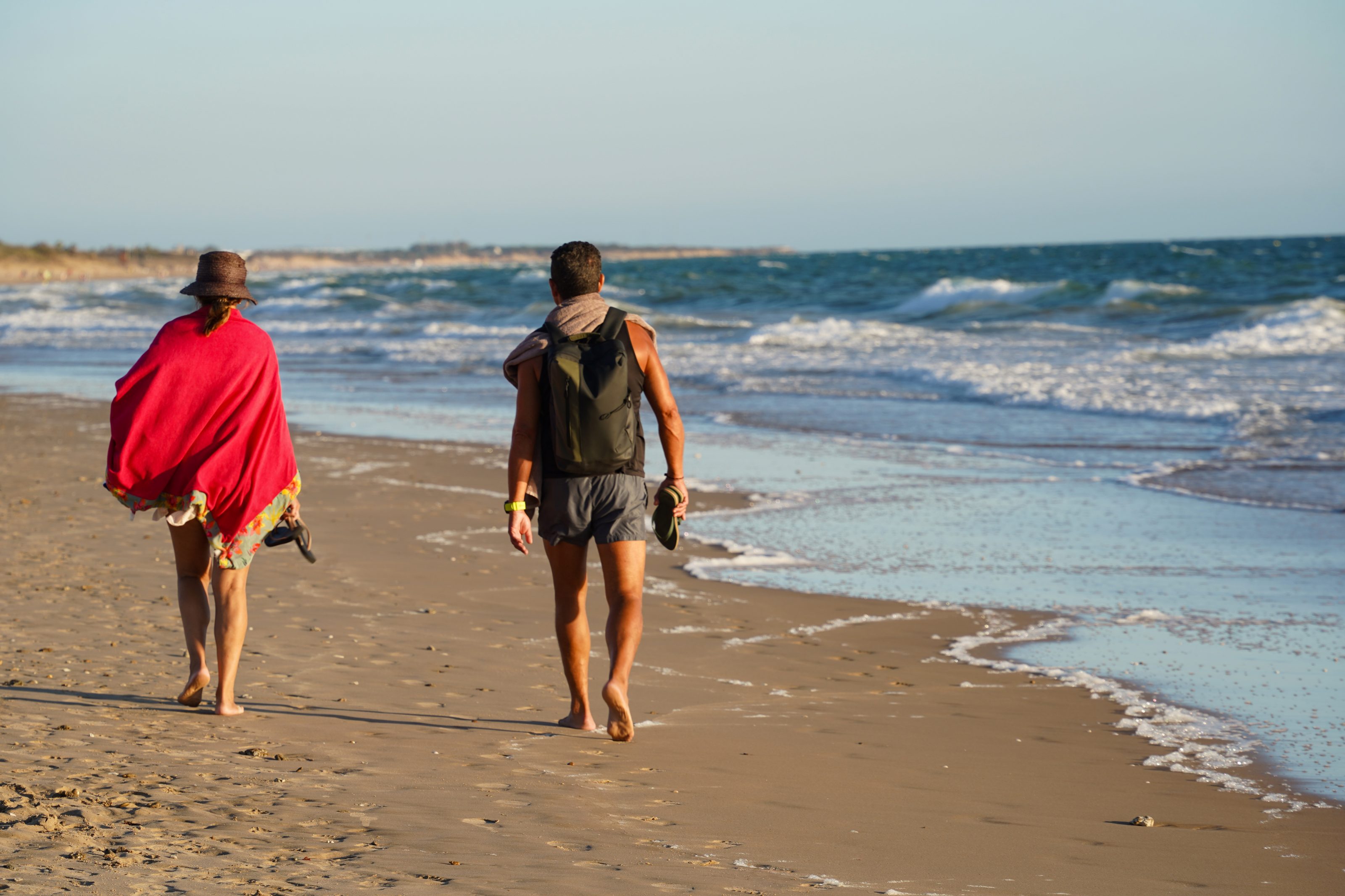 Two people walking barefoot along a sandy beach, with one wearing a hat and red shawl, while the other carries a backpack and sandals. The ocean waves gently lap the shore under a clear sky.