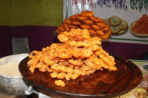 A close-up view of piles of  jalebi sweets arranged on a wooden tray, with a metal pot containing batter nearby, displayed at a food stall.