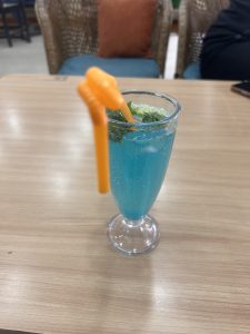 A clear glass filled with a blue beverage, garnished with mint leaves and ice, on a wooden table. There is an orange straw in the glass. The background shows a wicker chair with a blue cushion and an orange pillow.