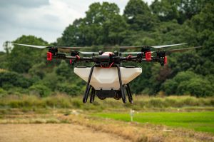 A drone flying over a grassy field with trees in the background, equipped with a tank and spraying mechanism likely for agricultural use.
