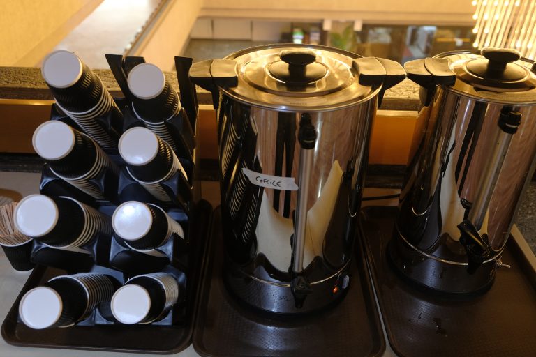 A coffee station with two large metal coffee urns on trays and a rack containing stacks of disposable coffee cups. A small container with stirrers is visible on the side.