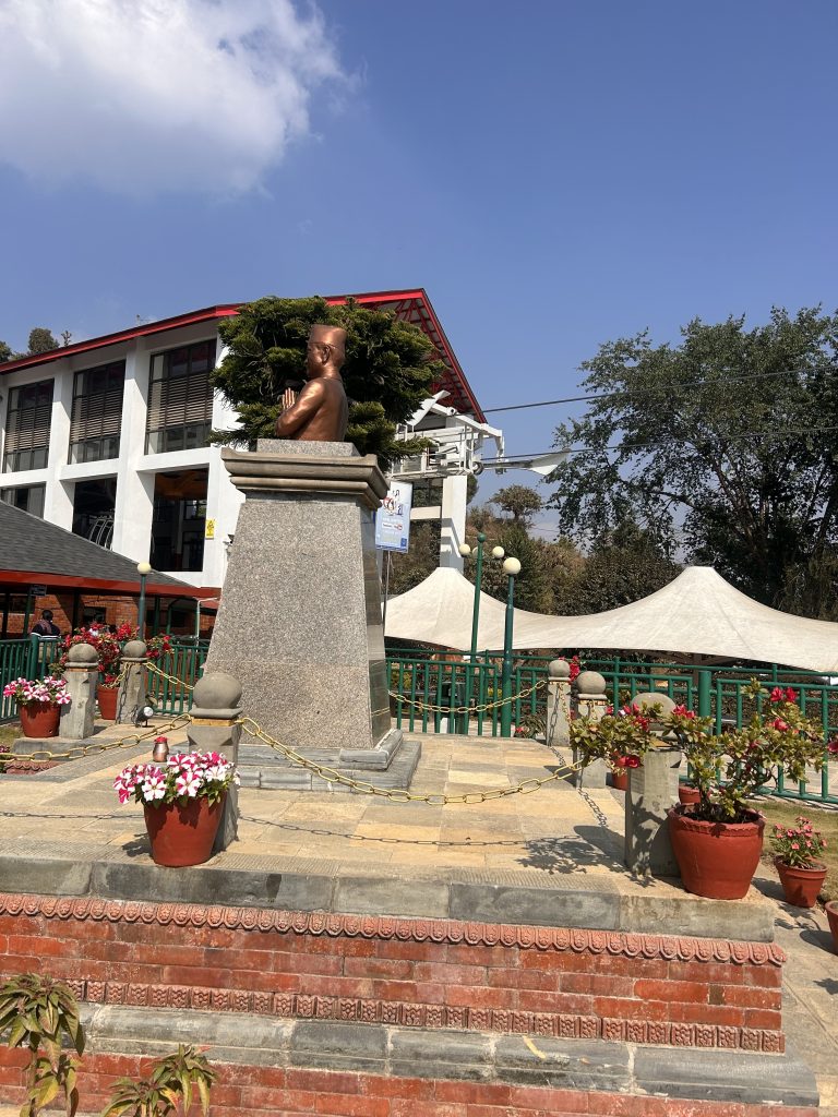 A statue of a man in a traditional pose stands on a pedestal outside, surrounded by potted plants and flowers.