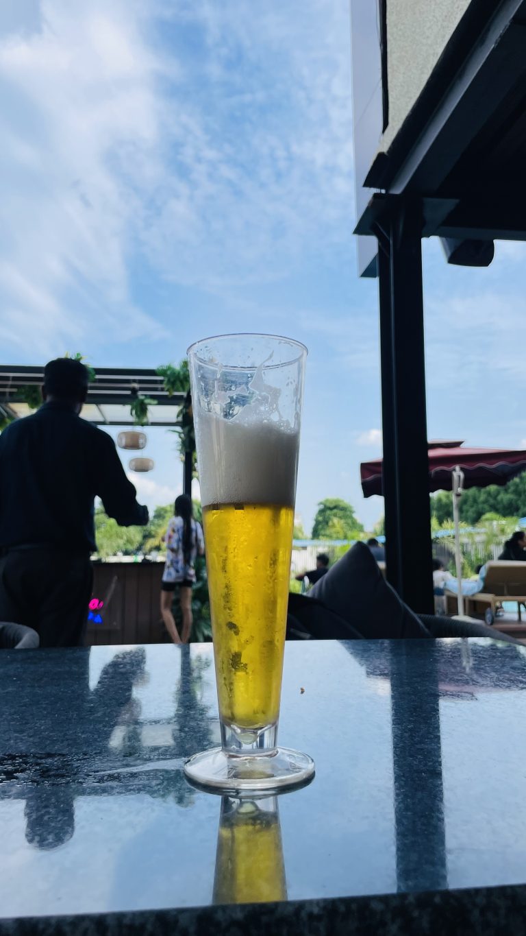 A tall glass of beer with a foamy head on a reflective table outdoors, with people and greenery in the background under a blue sky with clouds.