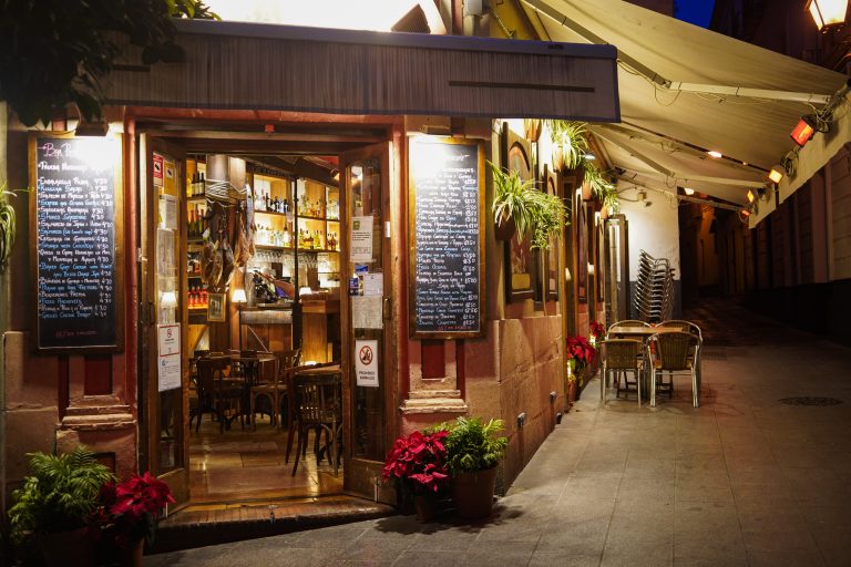 Exterior of a cozy bar or cafe at night with two chalkboards displaying menus on either side of the entrance. Inside, wooden tables and chairs are visible with shelves of bottles and hanging cured meats.