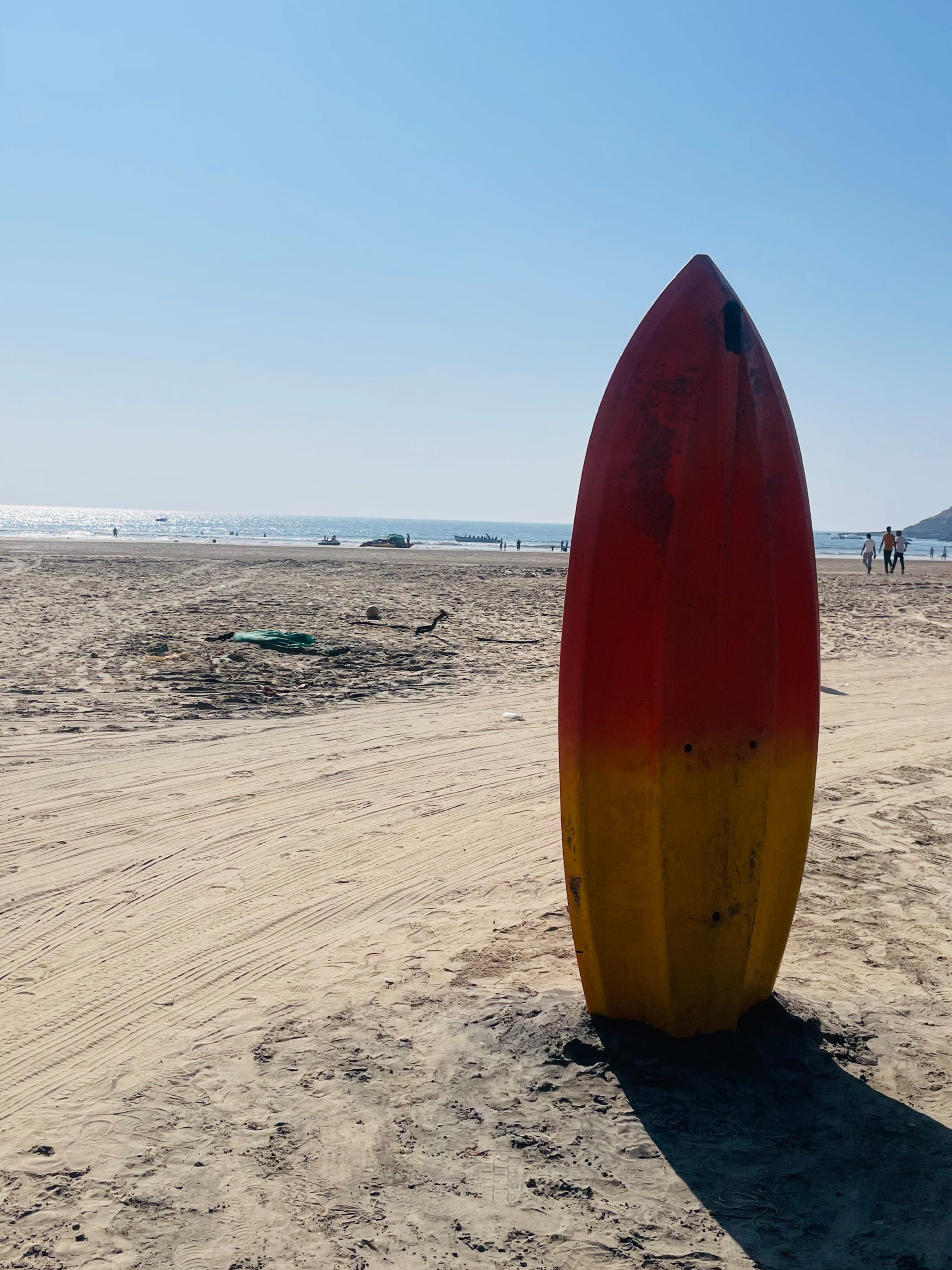 A colorful kayak standing upright in the sand on a sunny beach, with a few people, boats, and the ocean in the background under a clear blue sky.