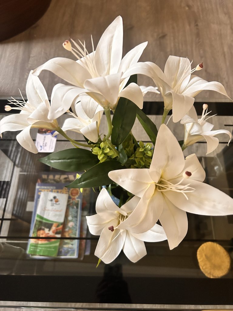 A bouquet of artificial white lilies with green leaves placed on a glass table. Below the flowers, there are magazines and a folded piece of paper visible through the glass surface.