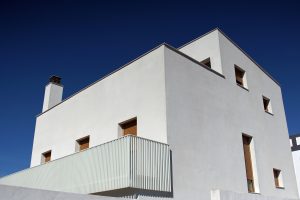 A modern, minimalist white house with rectangular windows and a chimney against a clear blue sky.