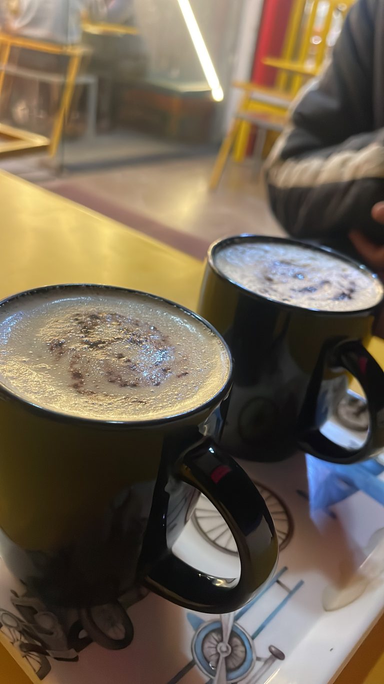 Two black mugs filled with frothy coffee placed on a table with a blurred background of a cafe interior.