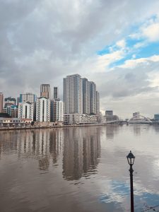 A scenic view of the Pasig River, where the waterway winds through the city, reflecting the urban skyline. Buildings rise along the banks, with boats occasionally passing by. The sky casts a warm glow, blending with the river’s ripples.