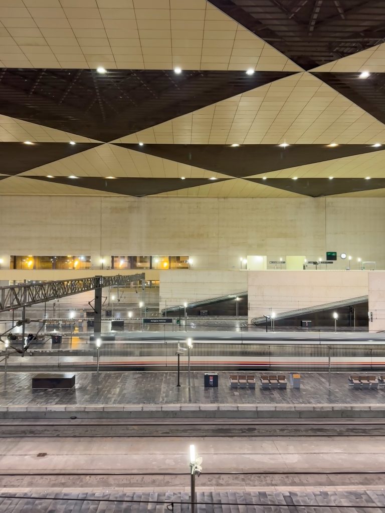 This is a nighttime view of Zaragoza-Delicias train station in Spain. The image captures the station’s modern architecture, featuring geometric ceiling panels with integrated lighting. A high-speed train is in motion on the platform, creating a blurred effect. The scene is illuminated by artificial lights, highlighting the sleek and spacious design