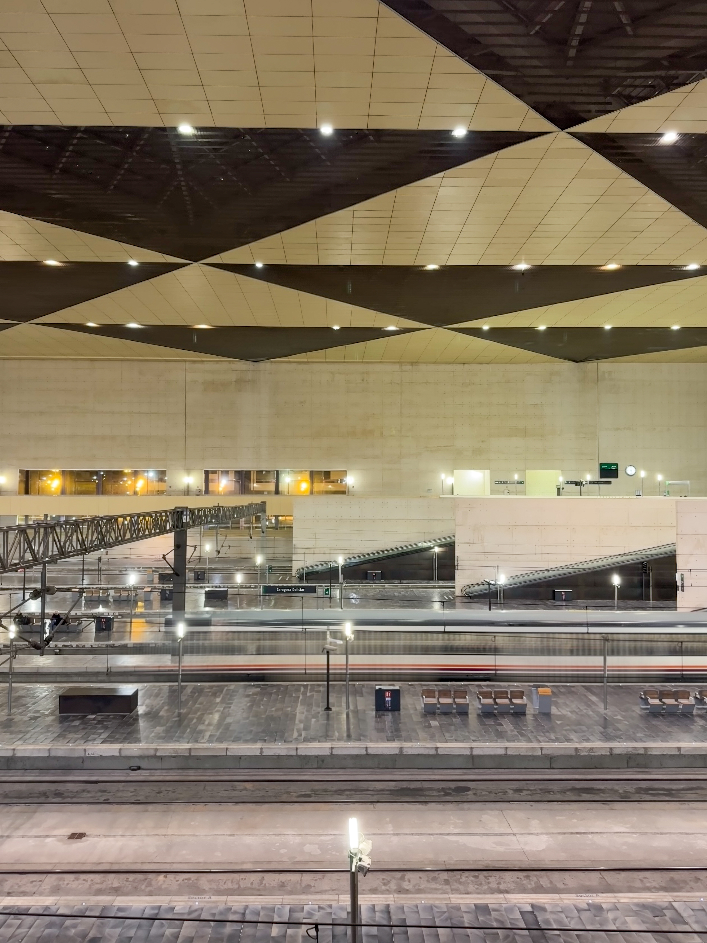 This is a nighttime view of Zaragoza-Delicias train station in Spain. The image captures the station's modern architecture, featuring geometric ceiling panels with integrated lighting. A high-speed train is in motion on the platform, creating a blurred effect. The scene is illuminated by artificial lights, highlighting the sleek and spacious design