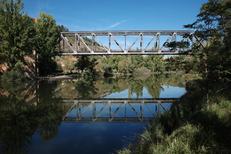 A metal train bridge over the Duero river in Soria, Spain. With trees on the banks and the reflection of the bridge and the blue sky.